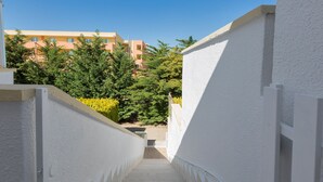 Una estrecha escalera de paredes blancas que conduce a la entrada de un edificio, rodeada de follaje y un cielo azul despejado.