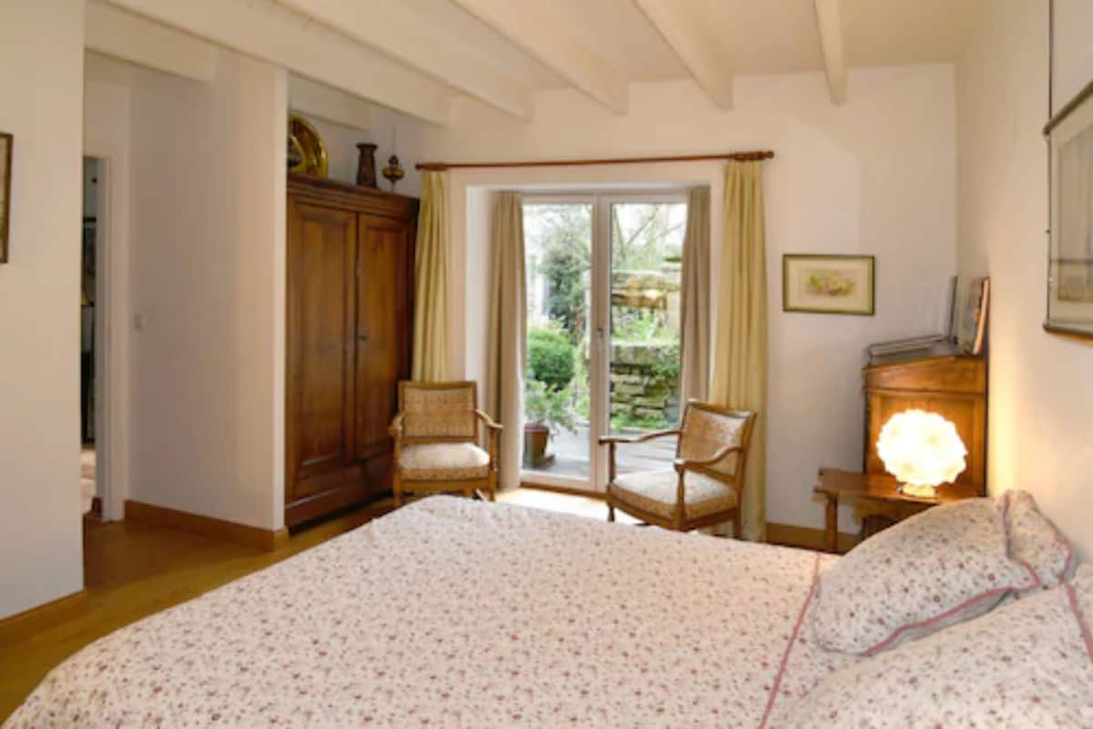 Een slaapkamer met een groot bed, twee stoelen, een houten kledingkast en een raam met gordijnen.
