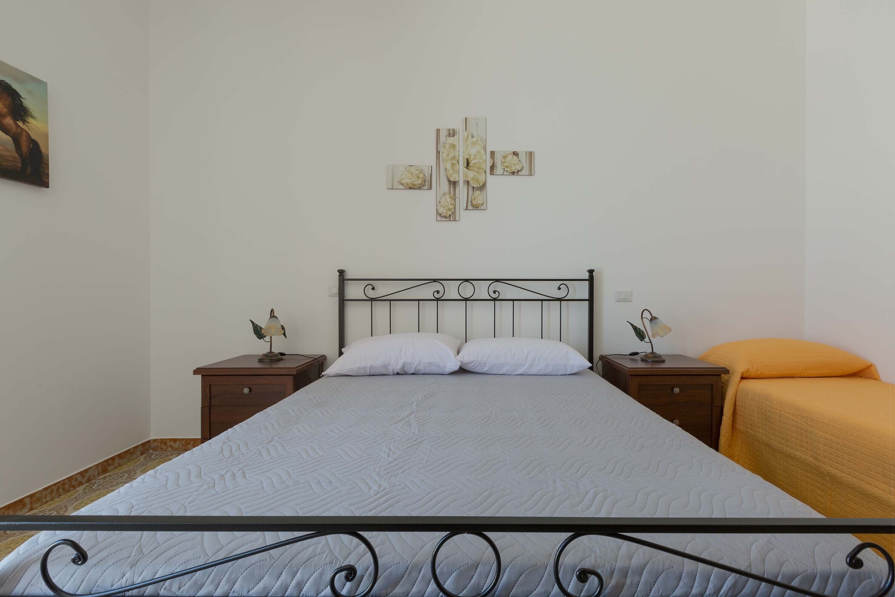 Een bed met metalen frame, twee nachtkastjes met lampen, een aan de muur bevestigd kunstwerk en een eenpersoonsbed met oranje deken.