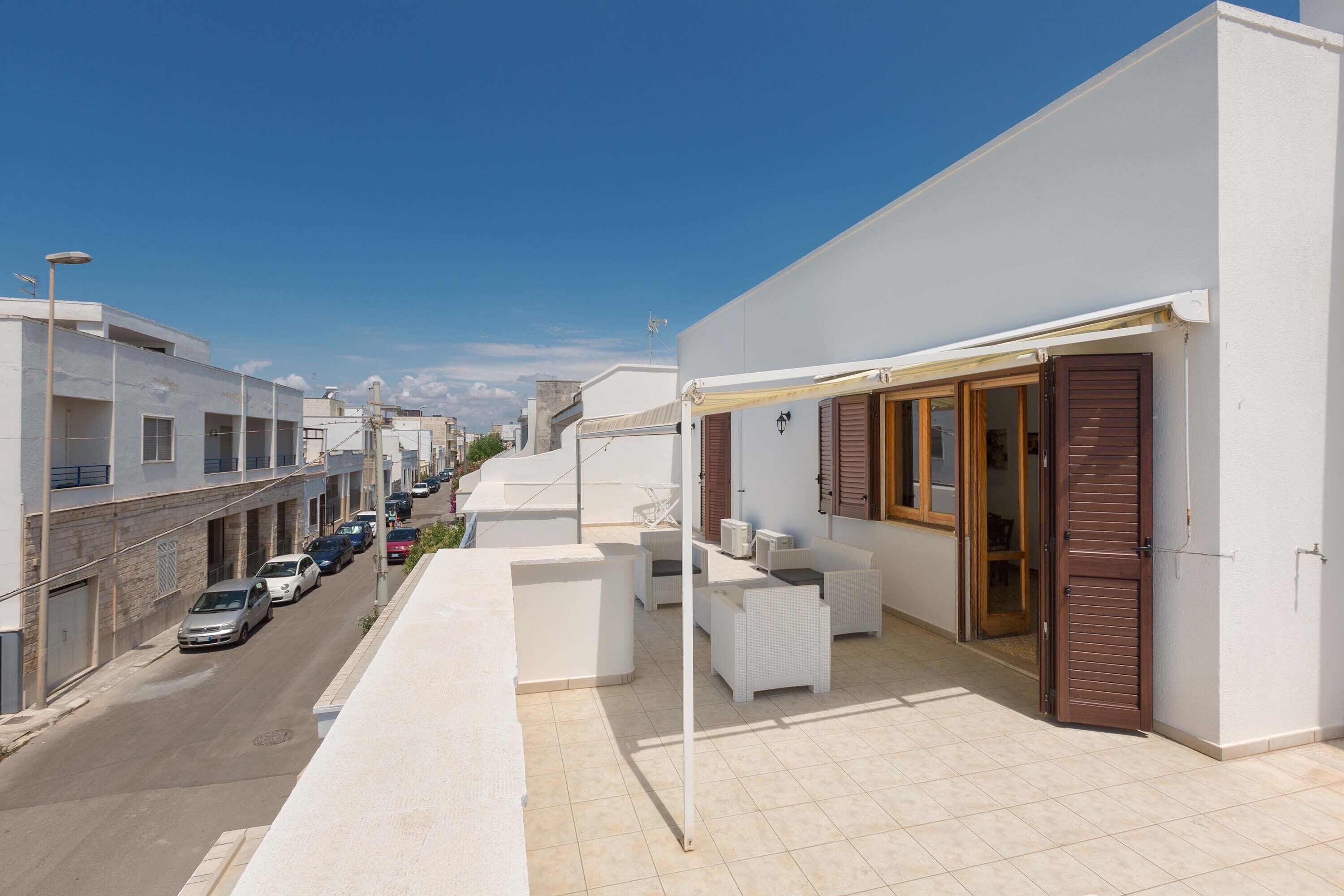 A residential street with parked cars, a white building with a balcony, and a clear blue sky.