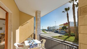 A balcony with a table and chairs, overlooking a street with palm trees and buildings. - Appartamento Sabbia Chiara (Porto Cesareo)