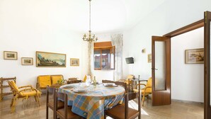 A dining area with a round table, chairs, and a chandelier. There are framed pictures on the walls and a window with curtains. - Appartamento Sabbia Chiara (Porto Cesareo)