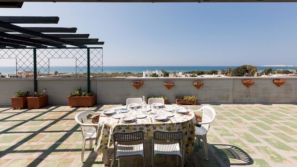 A rooftop terrace with a table set for dining, potted plants, and a view of the sea. - Attico La Baia (Gallipoli)