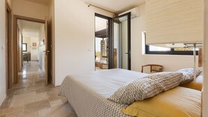 A bedroom with a bed, a window with blinds, a door leading to another room, and a hallway.