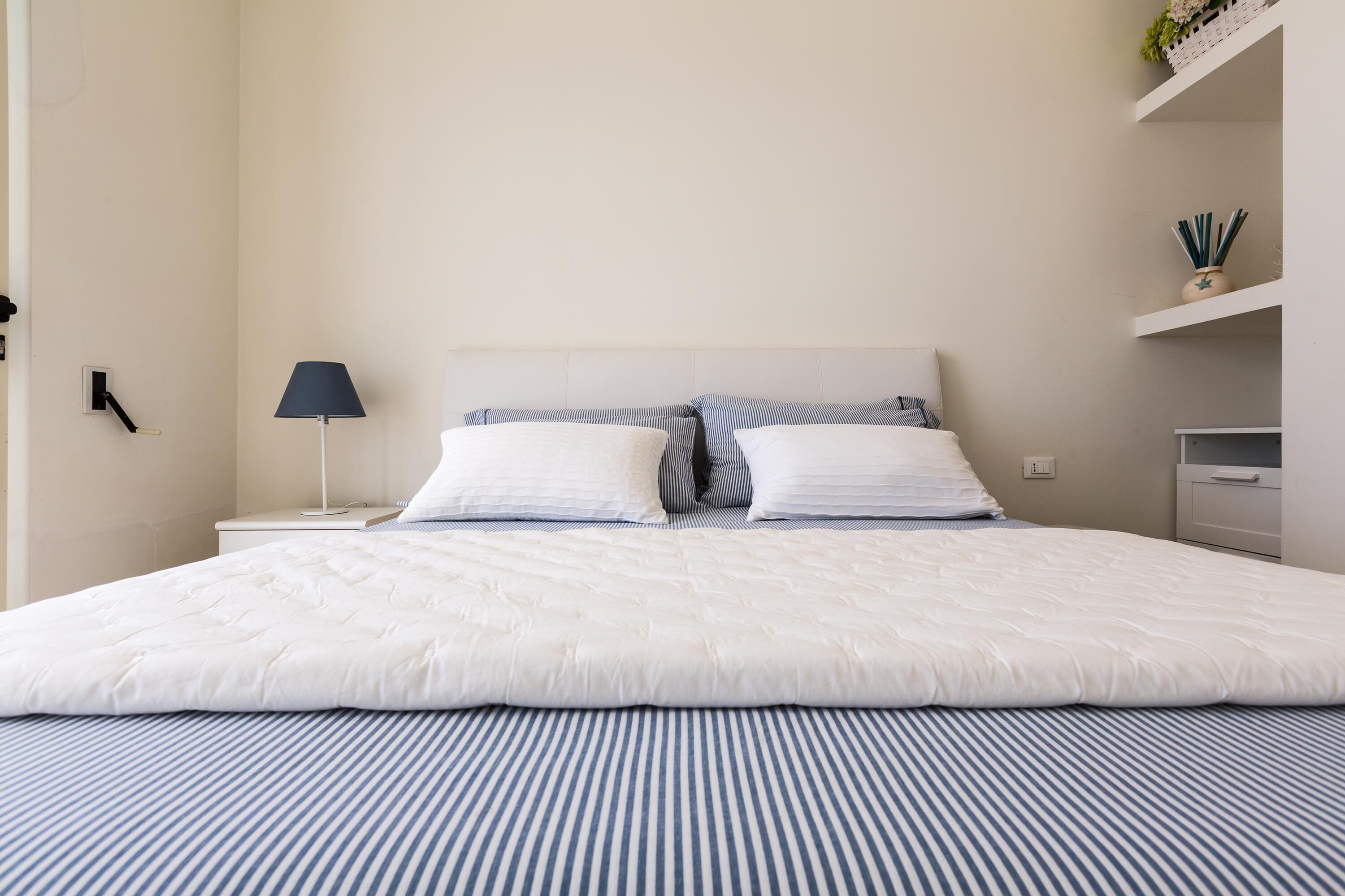 Una cama bien hecha con ropa de cama a rayas blancas y azules, una lámpara de noche y un pequeño estante con objetos decorativos.