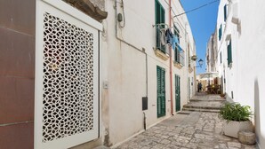 A narrow cobblestone street flanked by white buildings with green shutters and a decorative white lattice on the left. - La Casa di Clarissa (Otranto)