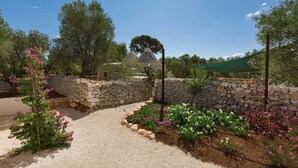 Un chemin de pierres mène à un bâtiment en pierre surmonté d’un toit conique, le tout étant entouré de plantes et d’arbres en fleurs.