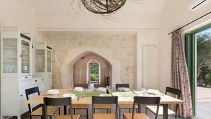 Un espace repas avec une grande table dressée pour six personnes, entourée de chaises, avec un mur en pierre et une vue sur la verdure à l’extérieur.