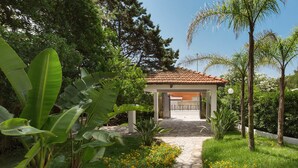 A covered outdoor area with a stone pathway leading to a building entrance, surrounded by lush greenery and flowering plants. - Villa Ariel (Porto Cesareo)