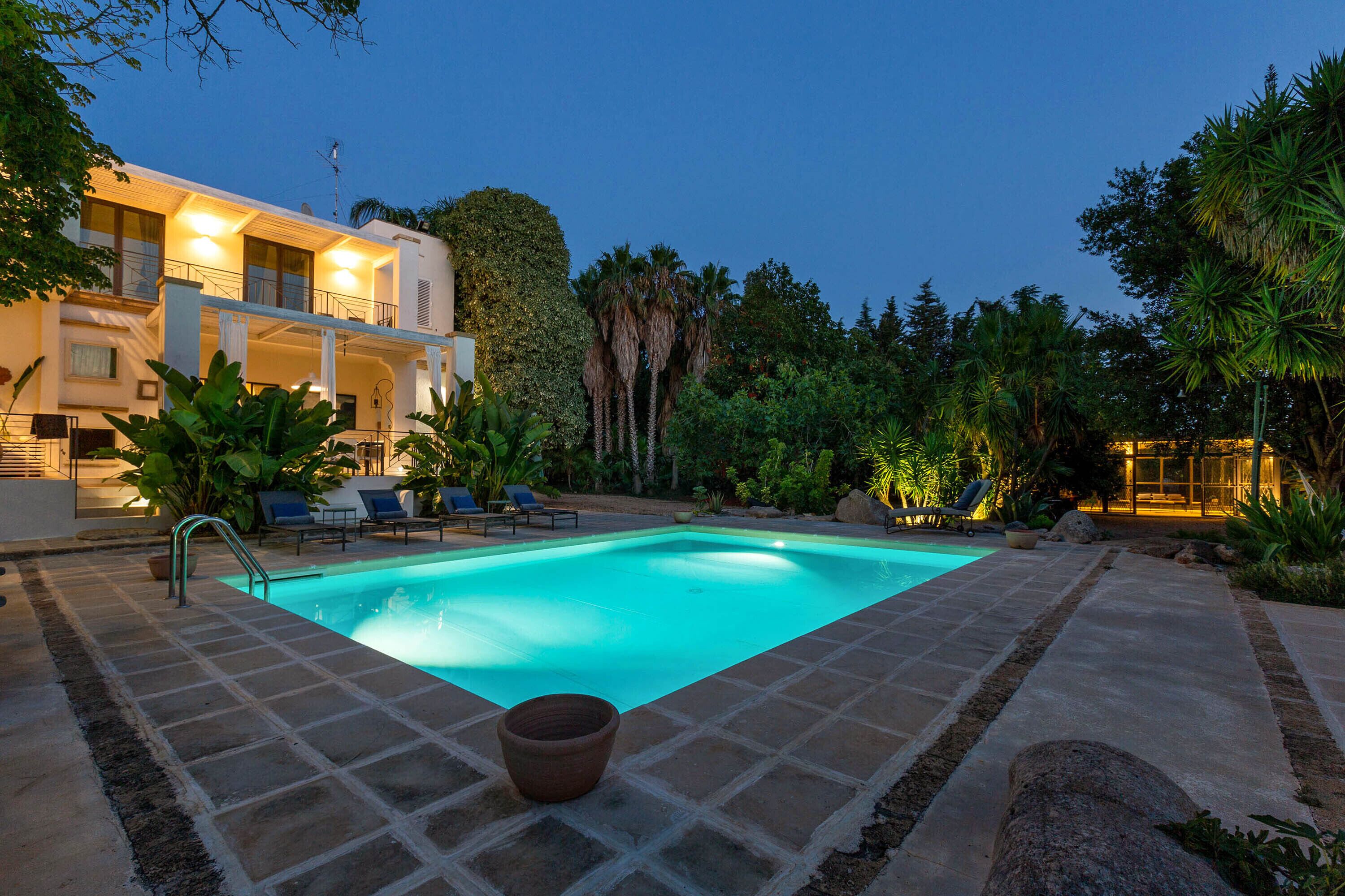 A swimming pool with a tiled deck, surrounded by lush greenery and a modern building with a balcony.