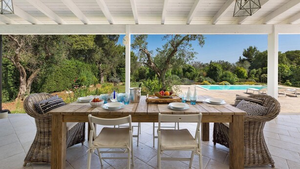 A covered outdoor dining area with a wooden table set for four, surrounded by wicker chairs, overlooking a pool and lush greenery. - Villa Chiara (Ruffano)