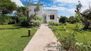 A white building with a stone pathway leading to the entrance, surrounded by greenery and flowering plants. - Villa Chiara (Ruffano)