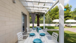 Un patio al aire libre con una mesa blanca lista para comer, rodeada de sillas blancas, y una pérgola arriba.