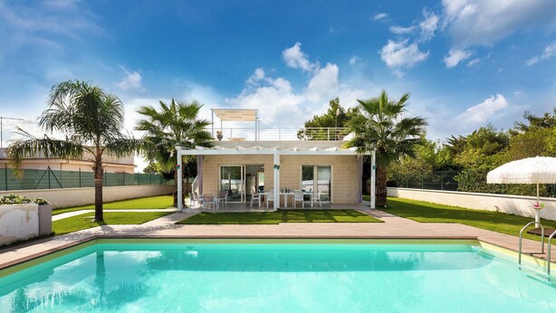 Una piscina con un área de terraza techada, palmeras y cielo despejado.