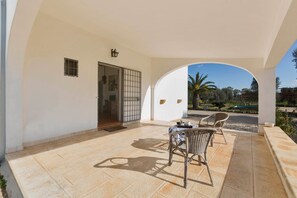A covered patio area with two wicker chairs and a table, a door with a metal gate, and a view of a swimming pool and greenery. - Villa Giardino dello Ionio (Nardò)
