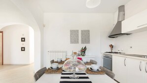 A modern kitchen with white cabinetry, a stainless steel range hood, and a dining area with a striped tablecloth and a glass cake stand. - Villa Hoara (Brindisi)