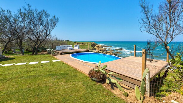 A swimming pool with a wooden deck, surrounded by a grassy lawn and cacti, overlooking the ocean. - Villa Hoara (Brindisi)