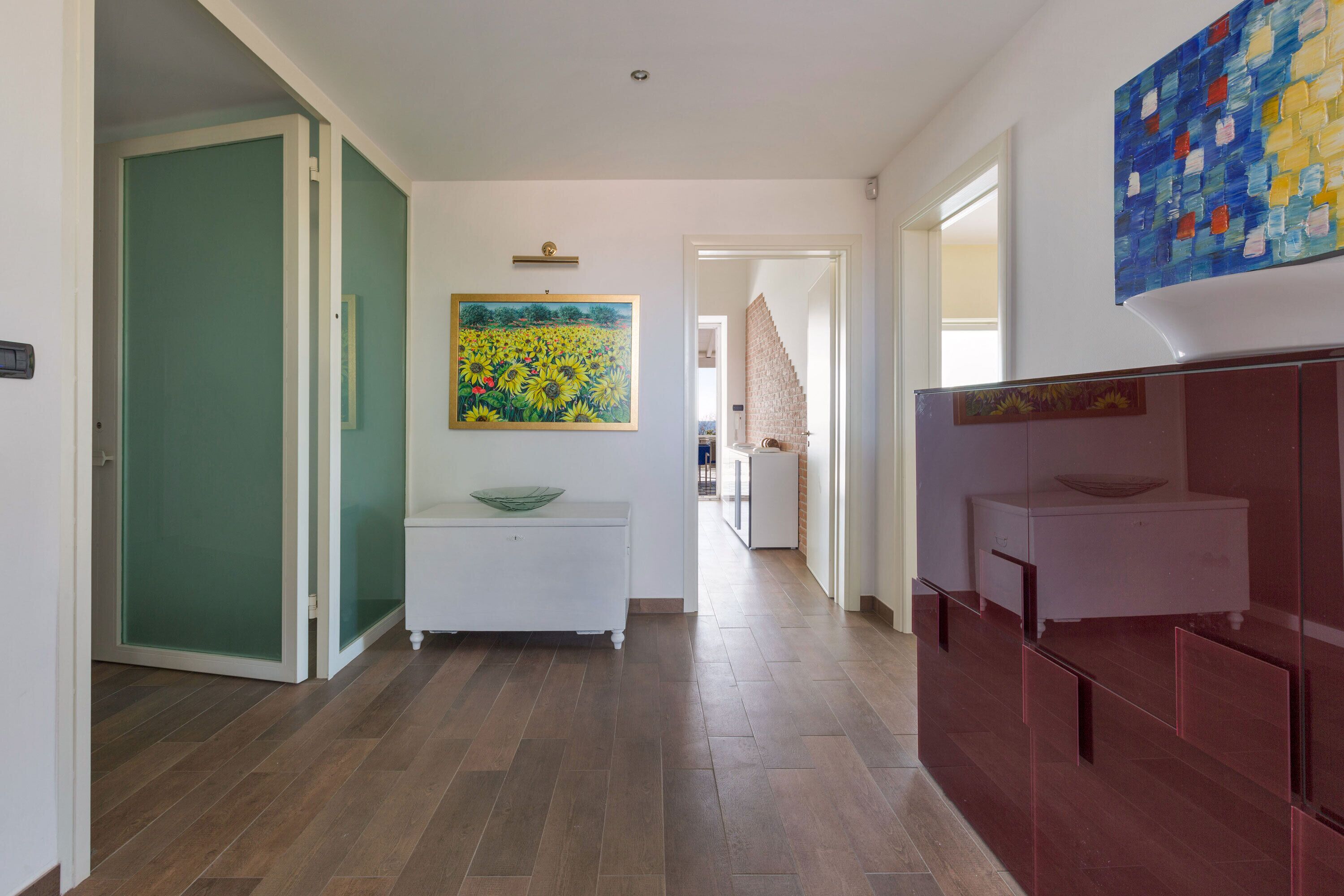 A hallway with a white bench, a painting, and a modern bathroom with a large mirror.