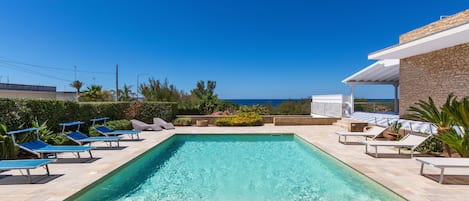 A rectangular swimming pool with clear blue water, surrounded by lounge chairs and a covered area with a view of the sea.