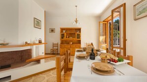 Un espace repas avec une longue table dressée pour le repas, des chaises en bois, une cheminée et, au fond, un coin cuisine.