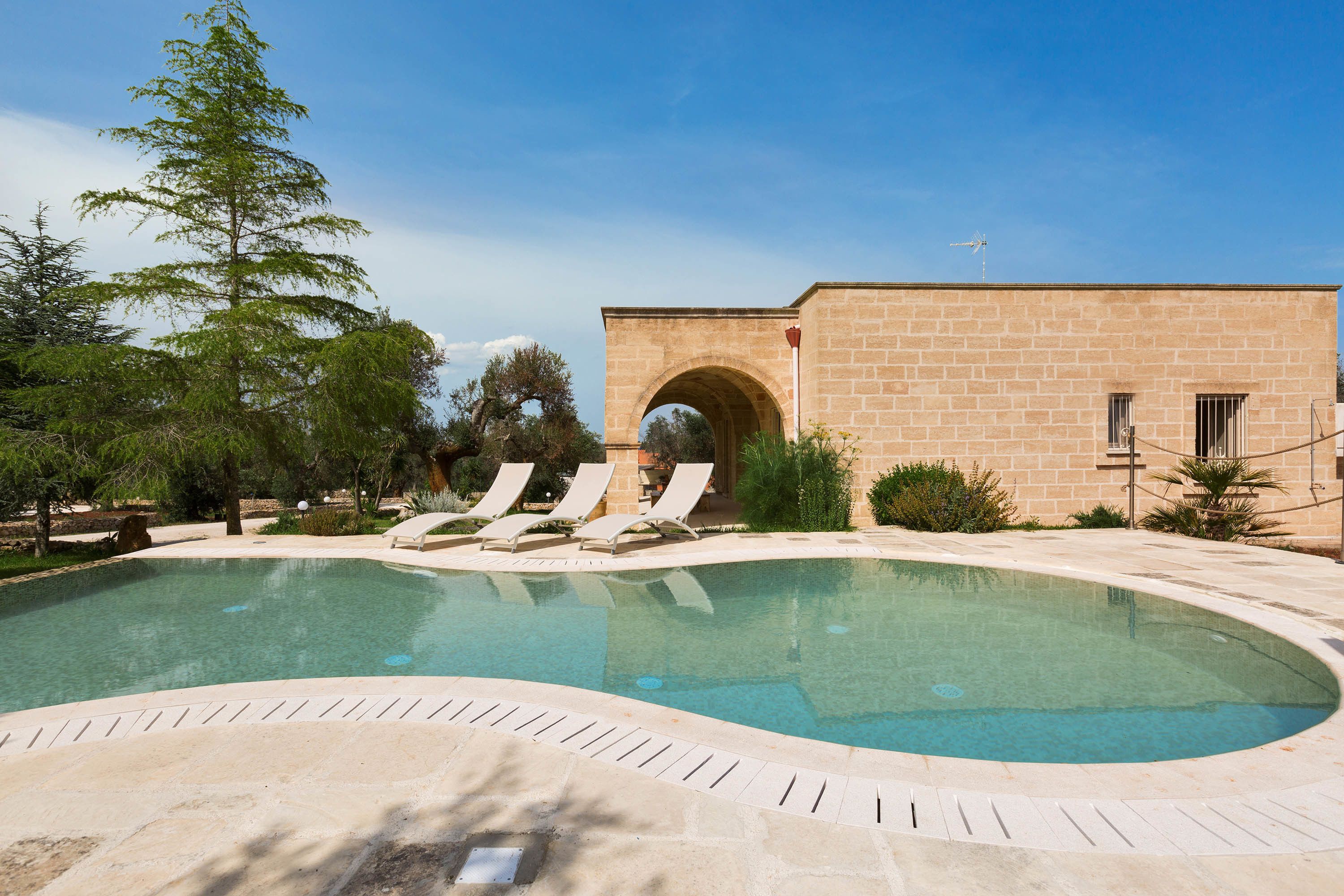 Une piscine entourée de chaises longues, un bâtiment en pierre doté d’une arche, et une végétation luxuriante.
