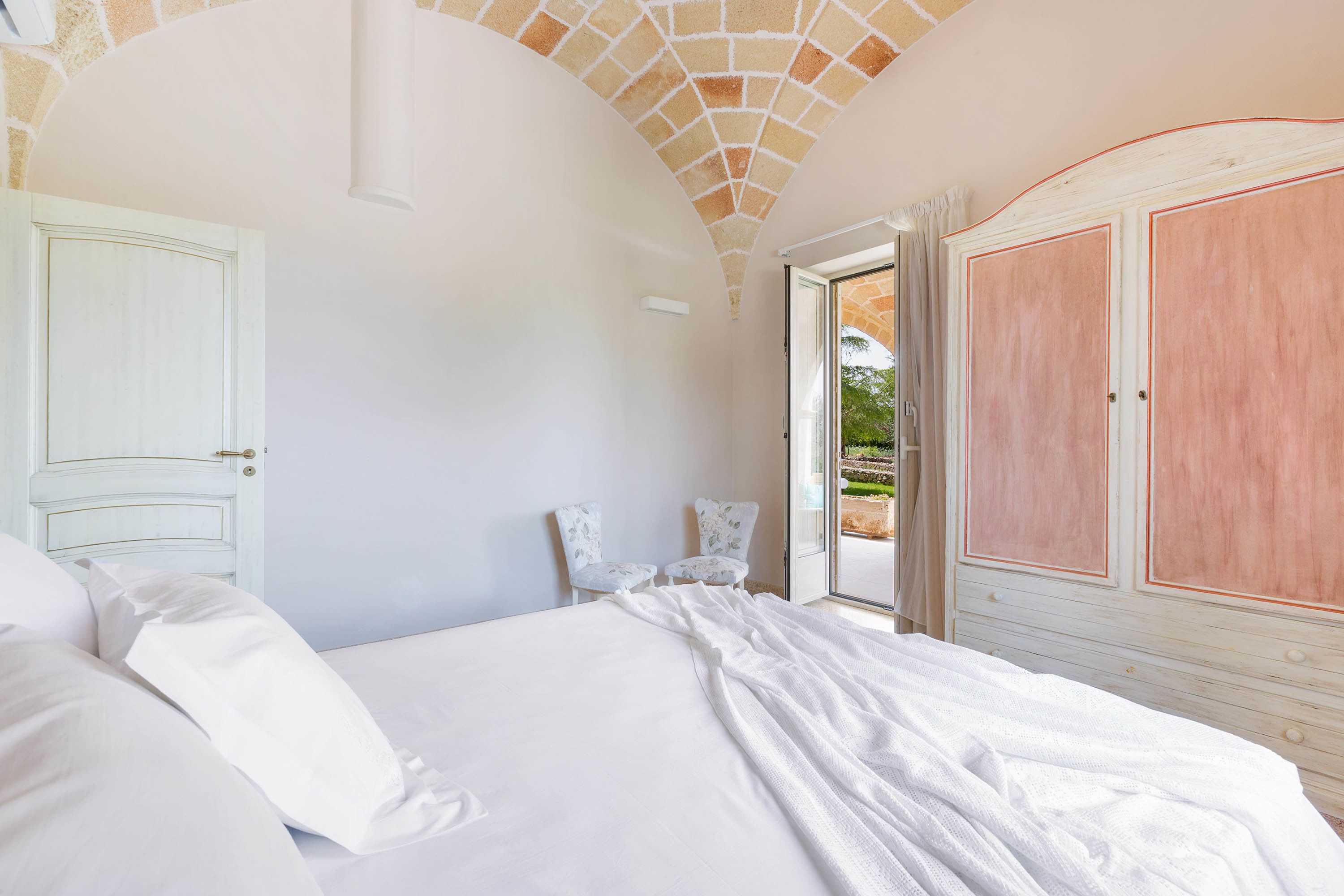 Une chambre à coucher avec un grand lit, du linge de lit blanc, une porte blanche et une vue sur l’extérieur par une porte ouverte.