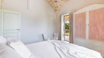 Une chambre à coucher avec un grand lit, du linge de lit blanc, une porte blanche et une vue sur l’extérieur par une porte ouverte.