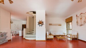 A spacious living area with a red-tiled floor, a central seating area with a sofa and chairs, a staircase leading to another level, and a wall-mounted mirror. - Villa Scirocco (Porto Cesareo)