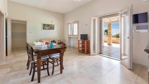 Un vaste espace de vie comprenant une table à manger, des chaises, un téléviseur posé sur un meuble et une porte-fenêtre coulissante donnant sur une terrasse extérieure.