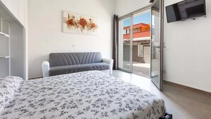 A bedroom with a bed, a grey sofa, a TV mounted on the wall, and a sliding glass door leading to an outdoor area.