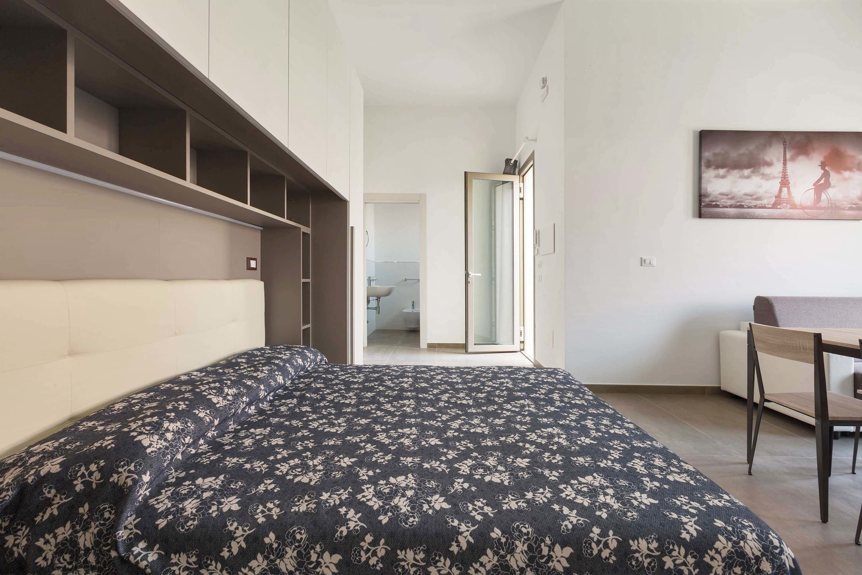 Ein Schlafzimmer mit einem Bett, einem Schreibtisch und einem Stuhl. An der Wand hängt ein Bild und eine Tür führt in einen weiteren Raum.