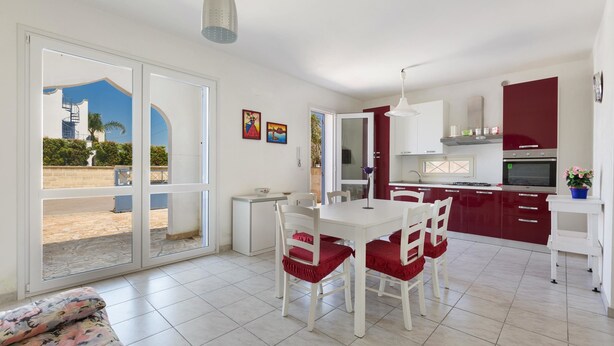Une cuisine moderne avec des meubles blancs et des touches de rouge, un coin repas avec des chaises et une table blanches, et une grande porte-fenêtre coulissante donnant sur un patio extérieur.