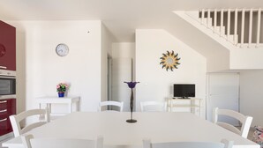 Un coin repas avec une table et des chaises blanches, une horloge au mur, un élément décoratif mural en forme de soleil, et un escalier menant à un niveau supérieur.