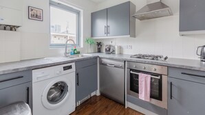 Ett modernt kök med gråa skåp, en inbyggd diskmaskin, en tvättmaskin, en spis och ett fönster som släpper in naturligt ljus.
