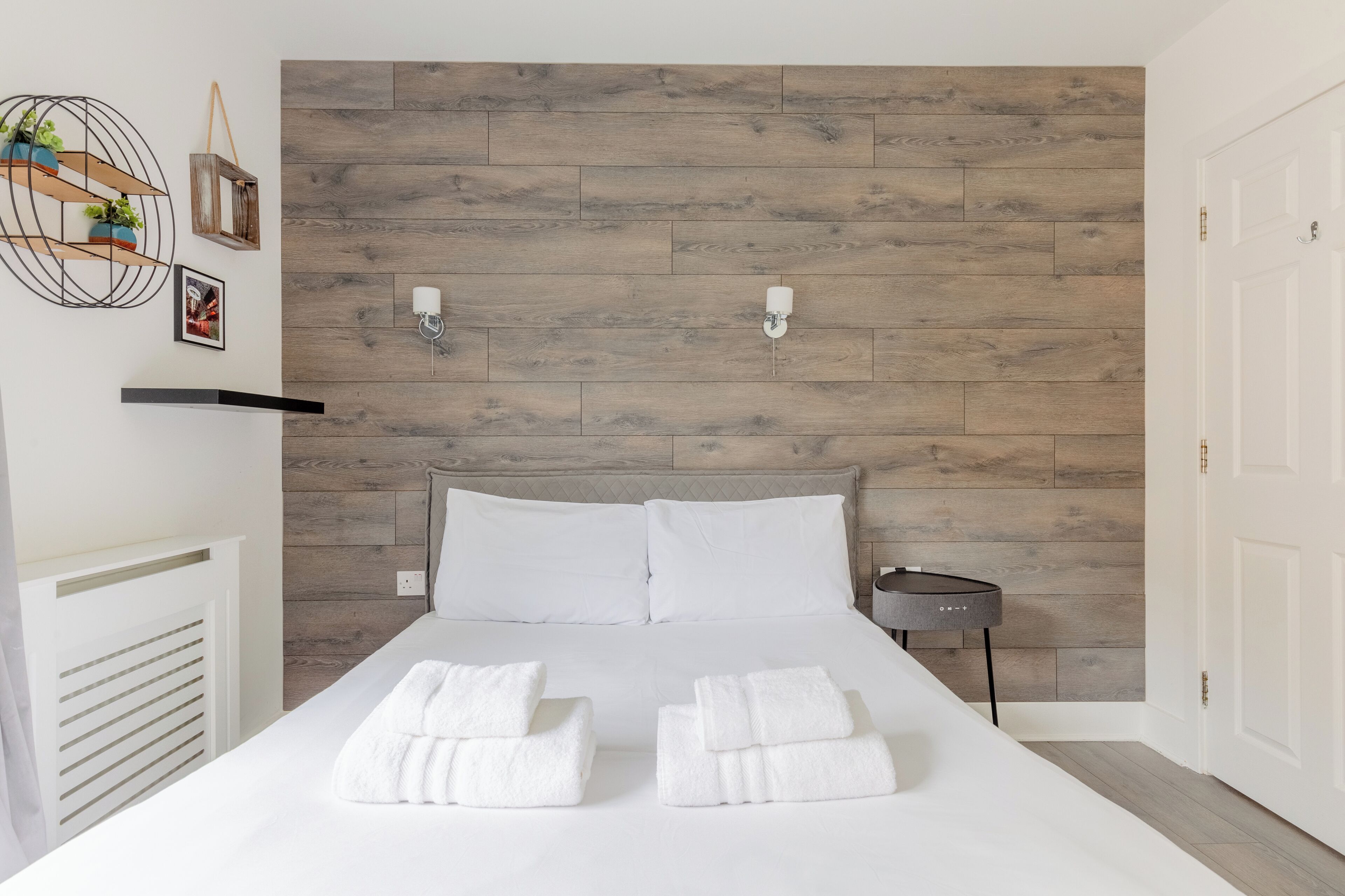 Una cama bien tendida con sábanas y toallas blancas, un cabecero de madera y una mesita de noche con una lámpara.