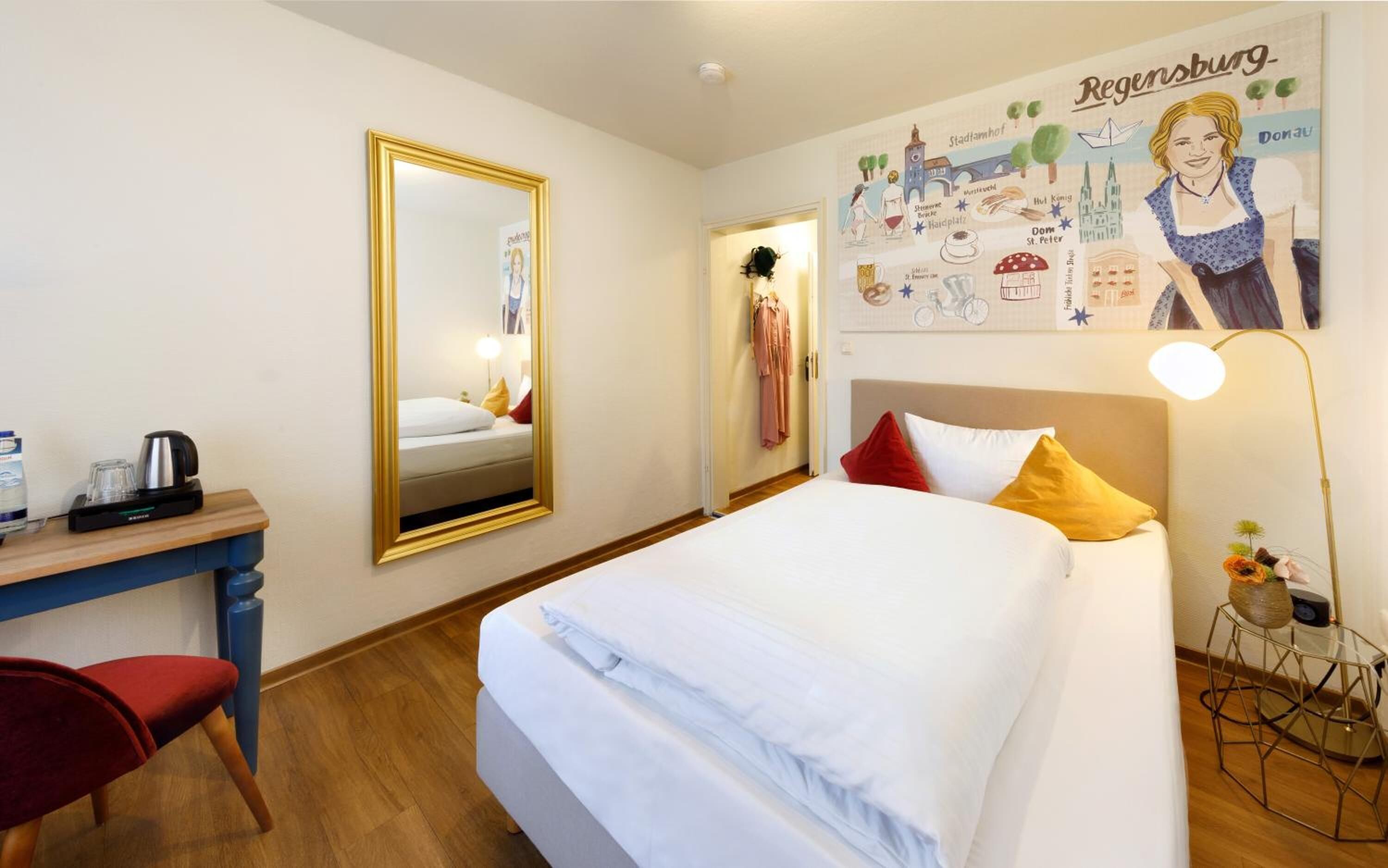 A hotel room with a bed, a desk, a chair, a mirror, and a wall with a cityscape illustration.