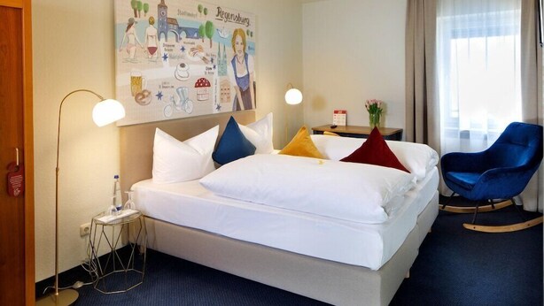 Ett hotellrum med en säng, ett skrivbord, en stol, en lampa och en affisch på väggen.