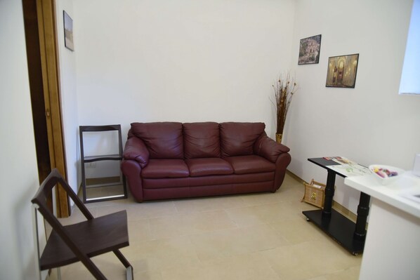A living room with a brown leather sofa, a wooden chair, a small table with a basket, and two framed pictures on the wall. - La Corte Di Ibla (Ragusa)