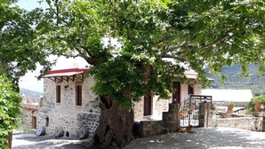 A stone building with a red roof, a large tree in front, and a view of a mountainous landscape. - Traditional house koukounara (kissamos)