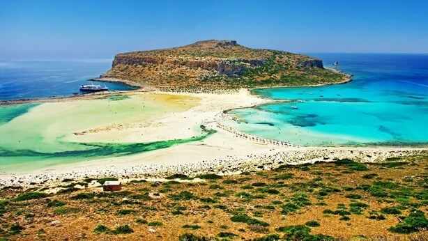 A coastal landscape with a small island, clear turquoise waters, and a boat docked near the shore. - Traditional house koukounara (kissamos)