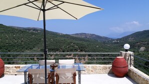 An outdoor dining area with a table and chairs, an umbrella, and a scenic view of green hills and mountains. - Traditional house koukounara (kissamos)