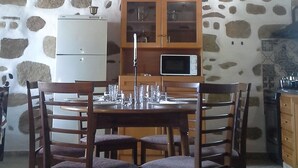 A rustic dining area with a stone wall, wooden ceiling beams, a wooden table with chairs, and a kitchen area with cabinets and appliances. - Traditional house koukounara (kissamos)