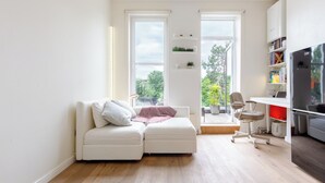 Ein modernes Wohnzimmer mit einem weißen Sofa, einem kleinen Schreibtisch und einem Sessel. Zwei Fenster bieten natürliches Licht und einen Blick ins Freie.