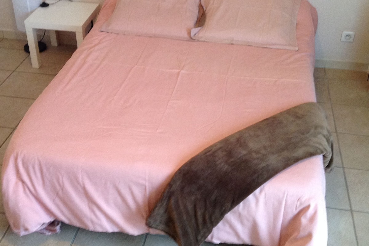 A bed with a pink bedspread and a brown blanket, a small white nightstand, and a white wall.