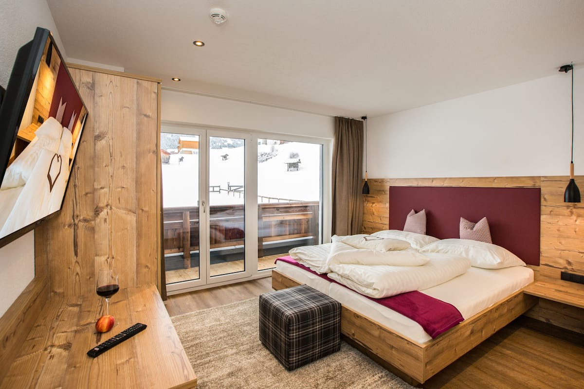 Ein modernes Schlafzimmer mit einem Holzkopfende, einem großen Bett mit weißer Bettwäsche, einem kleinen Nachttisch mit Fernbedienung und einer Glastür zum Balkon.