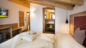 A modern hotel room with a wooden ceiling, a bed with white bedding, a small bench, a TV mounted on the wall, and a bathroom visible through an open door. - Alpinium (Fiss)