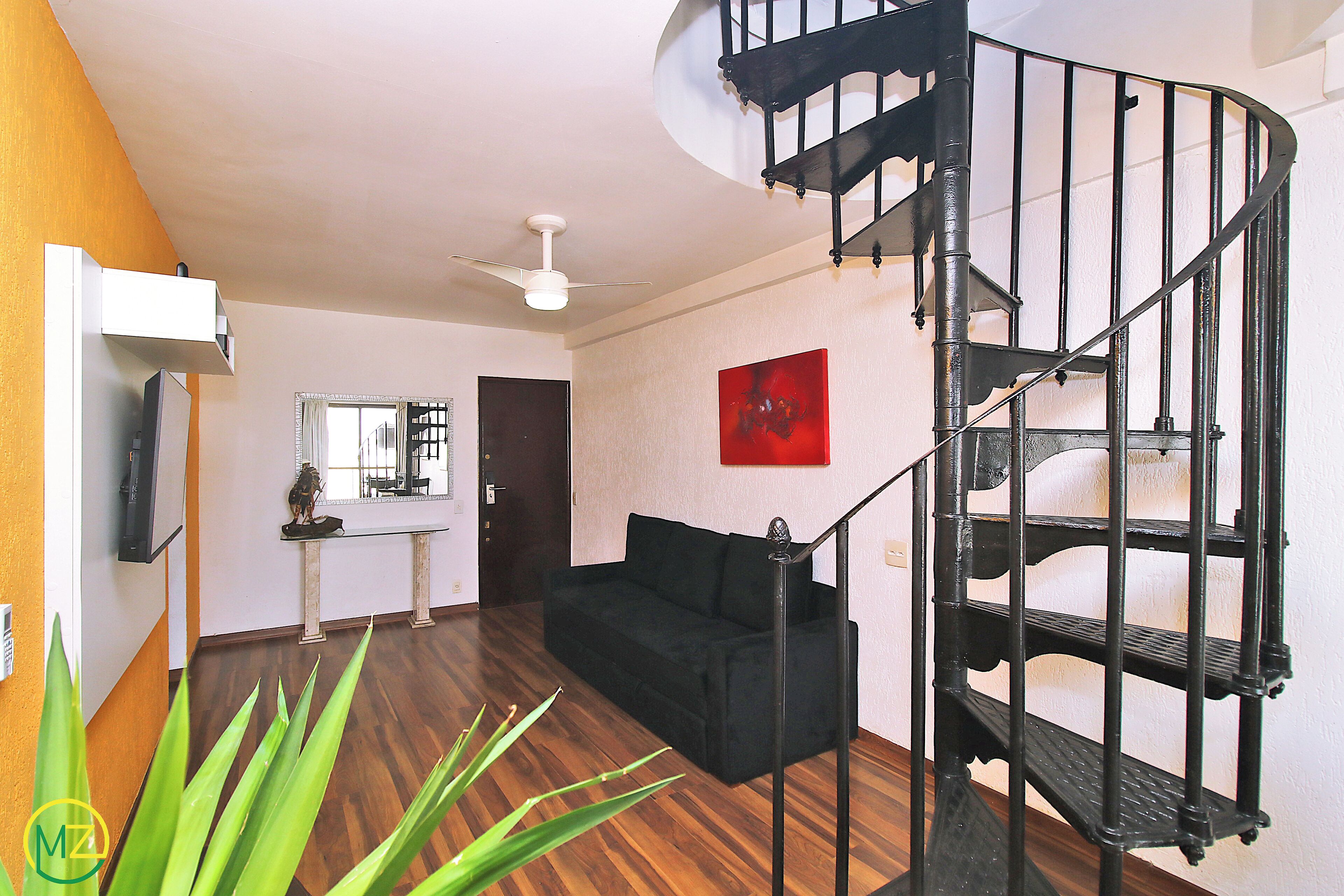 Uma sala de estar com sofá preto, escada, ventilador de teto e um quadro vermelho na parede.
