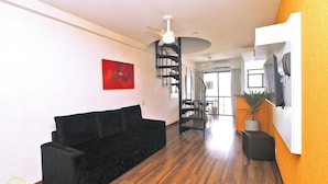 A living room with a black sofa, wooden flooring, a ceiling fan, a staircase, and a potted plant.