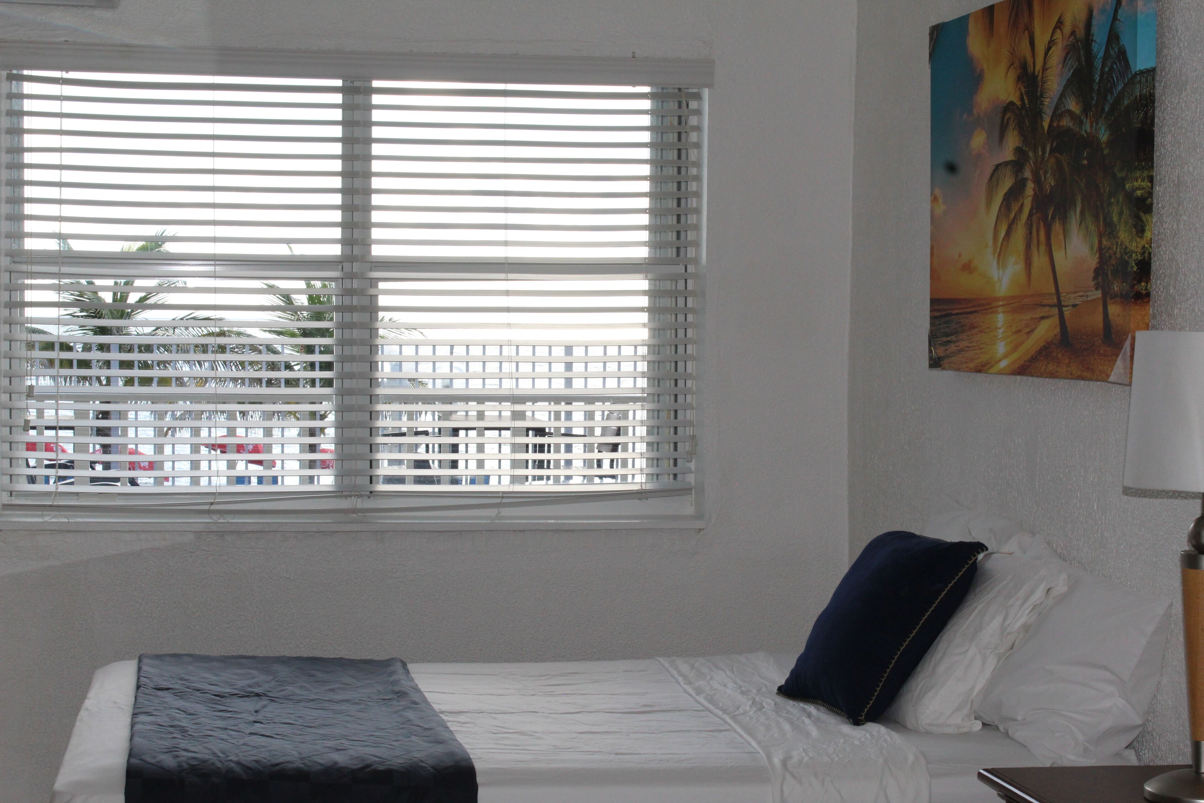 Ein Schlafzimmer mit Bett, Fenster mit Jalousien, einem Bild an der Wand und einer Lampe.