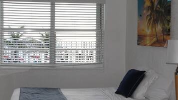 Ein Schlafzimmer mit Bett, Fenster mit Jalousien, einem Bild an der Wand und einer Lampe.